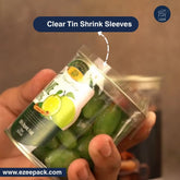 A hand holds a clear tin can with green contents and a printed label, demonstrating clear tin shrink sleeves. A dark blue text banner at the top reads 'Clear Tin Shrink Sleeves' with a dashed arrow pointing to the sealed top of the can.