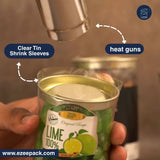 A hand holds a clear-labeled lime-flavored can while a metal heat gun heats the top. Text callouts identify clear shrink sleeves and heat guns, with a website watermark at the bottom.